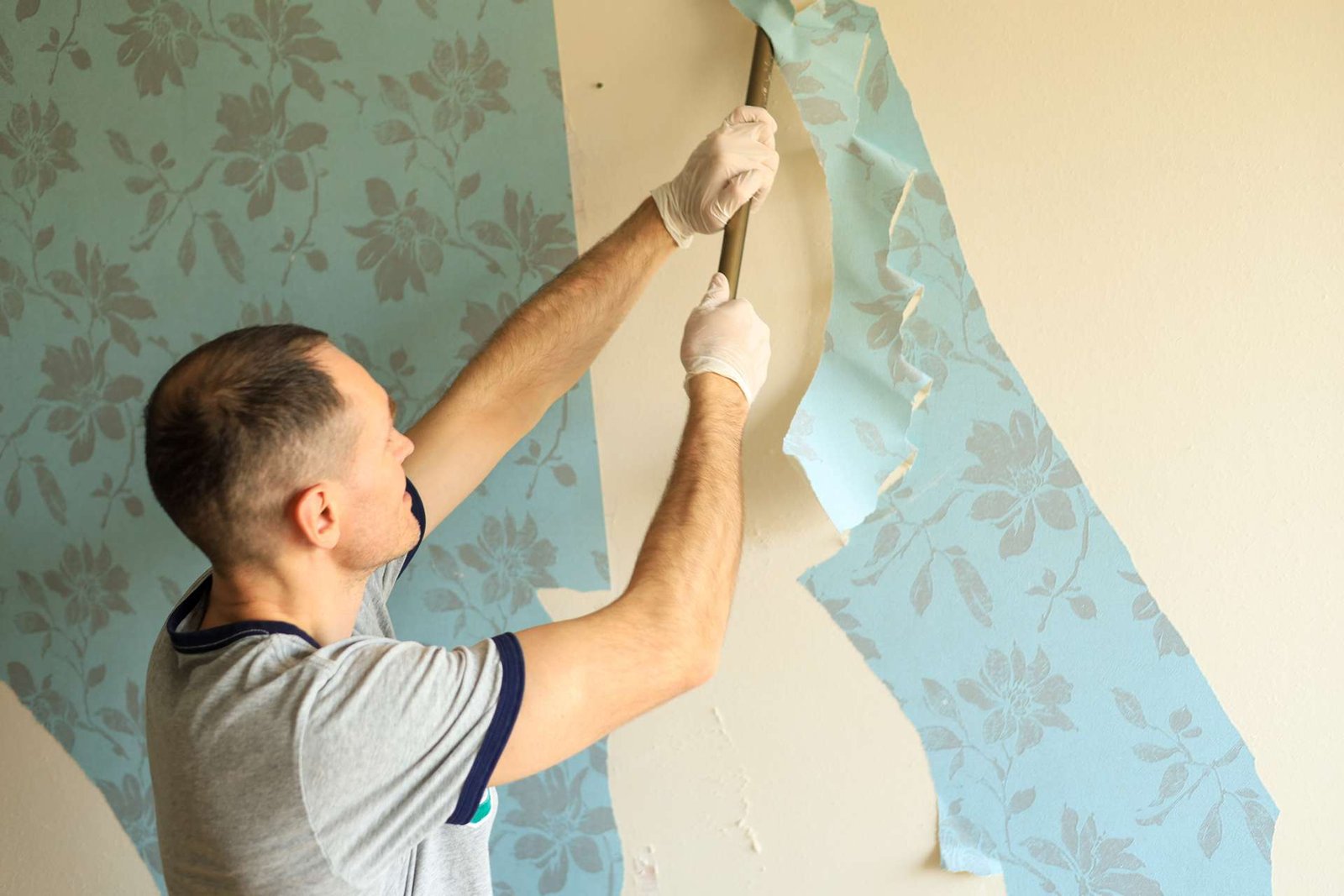 A professional uses a tool to remove blue floral wallpaper from an interior wall