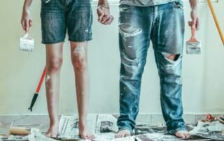 A waist-down view of a paint-splattered couple holding hands and surrounded by newspaper, white paint, and painting tools