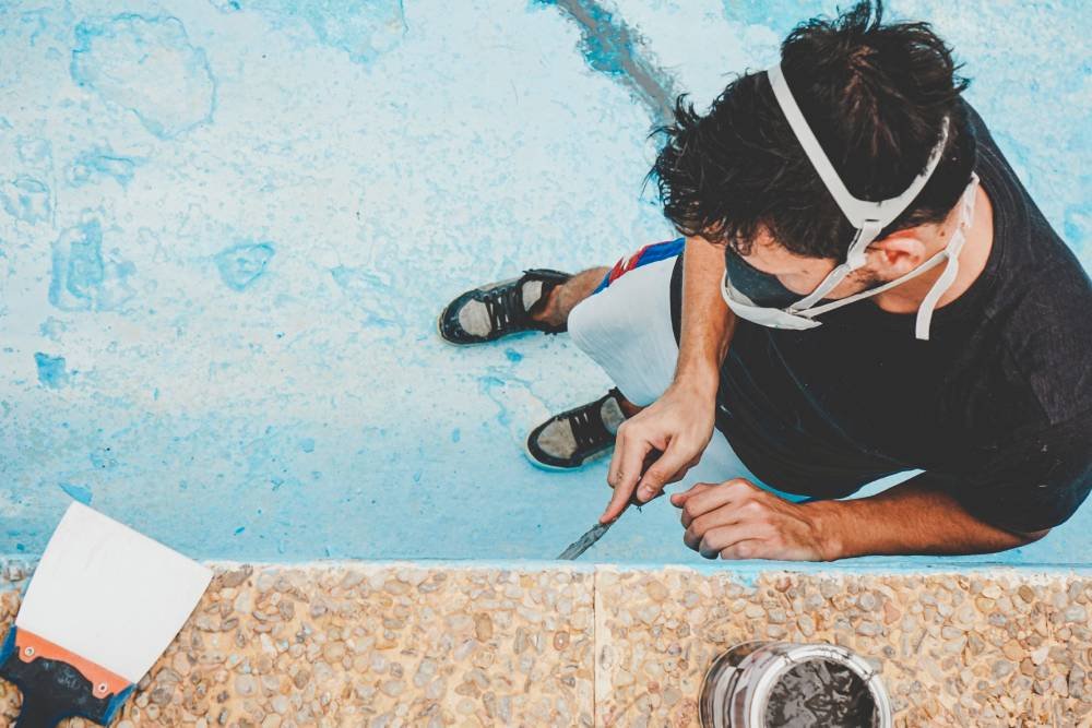 A man in a respirator mask scrapes a pool wall