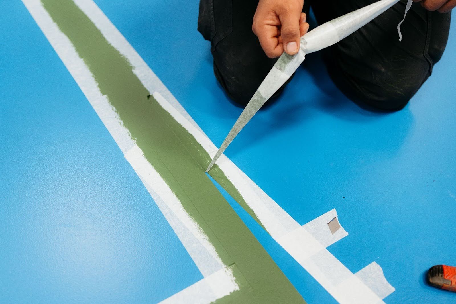 A professional removes masking tape surrounding a green stripe on a mostly blue floor